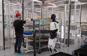 A Tesla Optimus humanoid robot walks through a factory with people. Predictable robot behavior requires priority-based control and a legal framework.